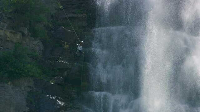Excited man rappelling down tall, rocky cliff next to beautiful waterfall - slow motion beauty