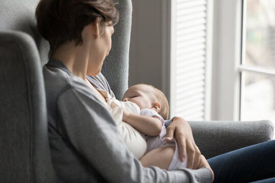 Happy days of motherhood. Peaceful woman young mother relax sit in armchair hold adorable baby child in arms watch with love care infant napping drinking breast milk feel close connection with child