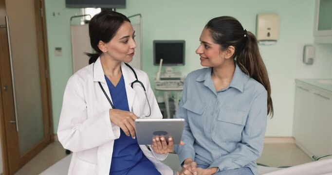 Happy diverse female doctor and patient talking in ultrasound examination room in clinic office, looking at scanning results on tablet, shaking hands with respect, gratitude, smiling