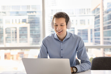 Positive handsome young business professional man in wireless headphones talking on video chat at laptop, using online conference call for work communication in city office, looking at screen, smiling