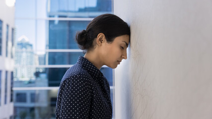 Stressed tired young Indian employee woman banging head against wall in burnout, feeling exhausted,...