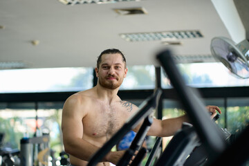 Sportsman naked shirtless smile and look at you for relax from body weighting with cardio machine in fitness gym.