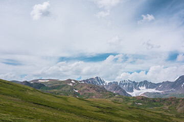 Fototapeta premium Scenic alpine landscape with green hills and rocks with view to large snow-capped mountain top and big glacier far away under clouds in blue sky in changeable weather. Awesome high snowy mountains.