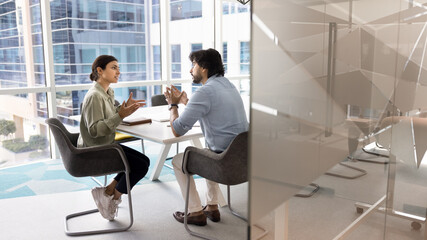 Two Indian business colleagues talking in meeting room behind glass wall, sitting at table...