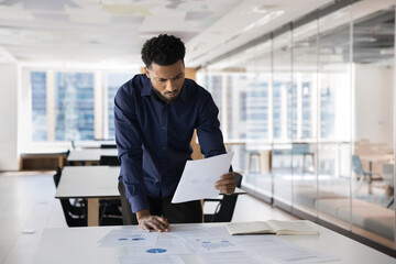 Focused concerned young African professional man reviewing marketing report at work table, reading document, frowning, leaning on desk with heap of papers, working in open office alone