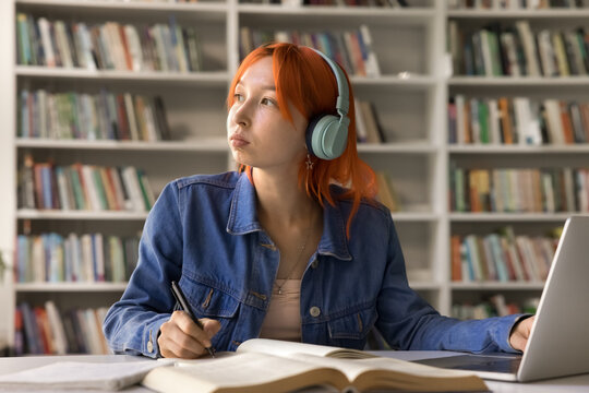 Pensive young woman student learn foreign language at online interactive video course sit by notebook screen in headset look aside think on text translation ponder on exercise before write to copybook