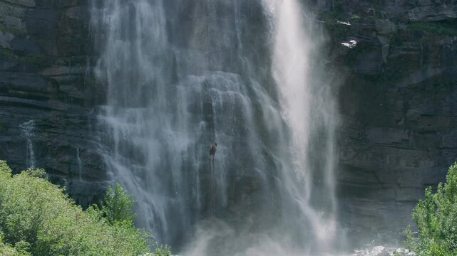 Wide shot of person rappelling down cliffside of large waterfall