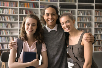 Diverse friends. Happy millennial male exchange student international university community member posing for portrait hugging shoulders two smiling young females groupmates in library after studying