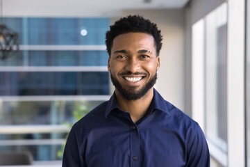 Cheerful handsome young African American businessman posing for head shot in office, looking at...
