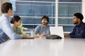 Fototapeta premium Confident Indian business team leader man holding corporate meeting at office table, talking to employees, sitting on bosses place at large conference desk, speaking, brainstorming