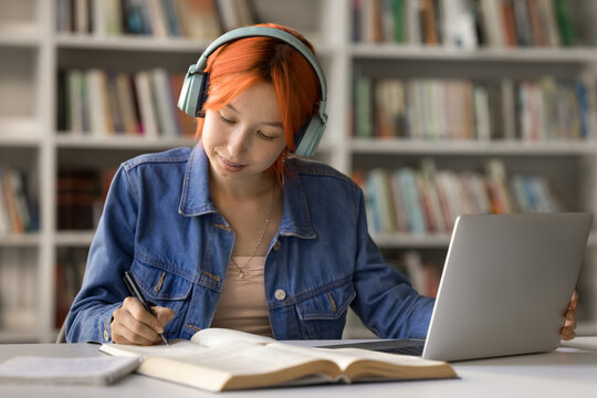 Hybrid communication. Smart young female remote class student in wireless headset attend online lecture sitting in library by notebook interact with teacher and group on distance take notes of webinar - Powered by Adobe