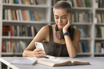 Smartphone in studying. Confident young hipster female student sit at desk in library hold phone look at screen text message search information online check task list at high school learning platform