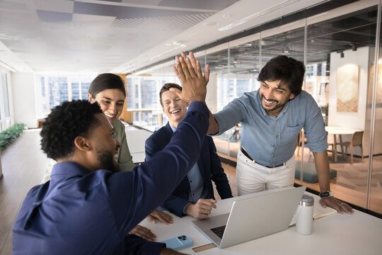Happy diverse young business friends giving successful high five at meeting in office, brainstorming on creative successful ideas, celebrating success achieving, clapping hands in win gesture