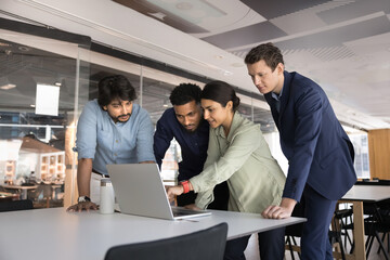 Multiethnic business team of young colleagues standing at laptop. Indian manager showing Internet application to coworkers, standing at computer with coworkers, pointing at screen, speaking