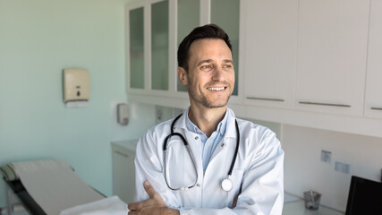 Positive thoughtful handsome male doctor posing in patient examination room in hospital, looking away with dreamy smile, standing with hands folded, thinking on medical career. Banner shot