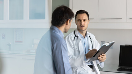 Serious doctor giving consultation to male patient, filling up survey, writing notes on pad,...