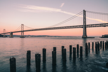 The Bay Bridge stretches over the water at sunset in San Francisco, with remnants of old wooden pilings in the foreground and hills in the distance.