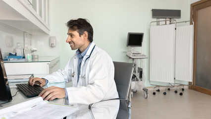 Happy middle aged Hispanic doctor man using desktop computer in clinic office, sitting at workplace table, typing on keyboard, smiling, enjoying online communication, medical Internet technology