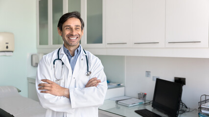 Cheerful successful Hispanic doctor man standing with arms crossed, keeping confident gesture, looking away with positive thoughts, smiling, laughing, thinking on success in medicine. Banner portrait