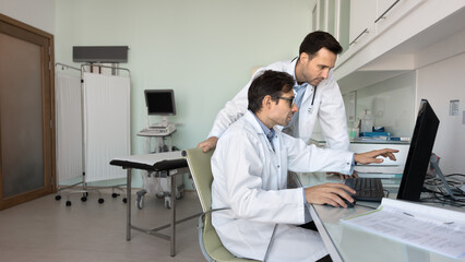 Obraz premium Two focused male doctors reviewing ultrasound examination result on computer monitor together, meeting at workplace table, discussing electronic medical report. Banner shot