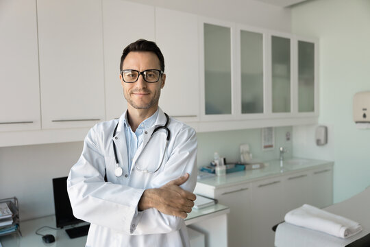 Positive confident young doctor man in glasses and white coat standing in clinic examination room with couch, looking at camera with arms folded, smiling, posing for medical professional portrait