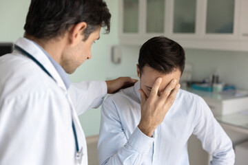 Fototapeta premium Shocked patient man getting bad news from doctor, learning serious hard diagnosis, covering face on couch in examination room. Medical specialist touching shoulder of unhappy depressed patient
