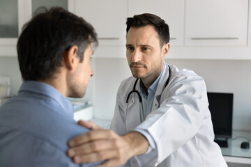 Serious caring doctor touching patients shoulder with sympathy, support, telling examinations bad...