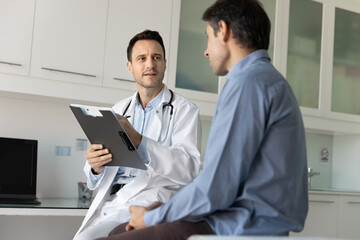 Caring young male doctor meeting with patient in clinic examination room, asking about symptoms,...