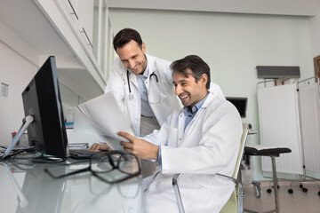 Fototapeta premium Two happy male doctors reading paper medical report at workplace with computer, working together in clinic examination room, reading and discussing document, smiling, laughing