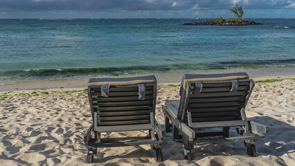 Two deck chairs with mattresses on the beach. Footprints on a sand. A tiny rocky island with a lone tree in the turquoise ocean. Clouds in the sky. Mauritius. Belle Mare.