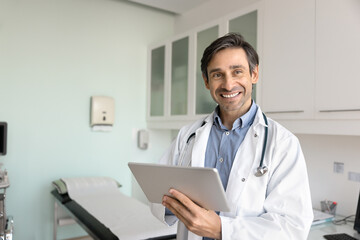 Happy Latin doctor man thinking on records on tablet, holding digital device, looking away, smiling, working on electronic data in patients examination room with couch