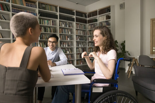 Learning equality. Smiling teen girl high schooler using wheelchair an inclusive class pupil student hold pleasant conversation share funny idea with laughing groupmates friends on workshop at library - Powered by Adobe