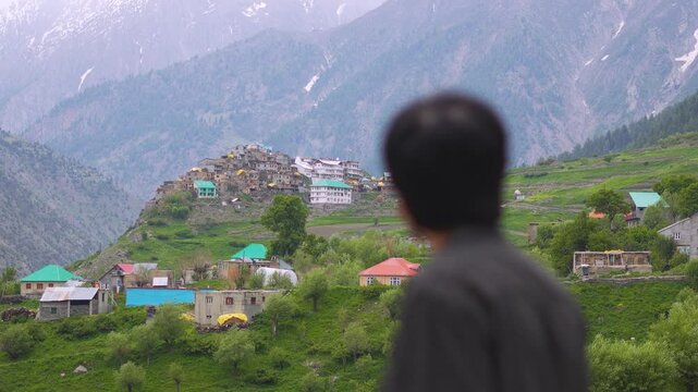 4K shot of a defocused Indian man looking at Triloknath village in front of the snowy Himalayan mountain peaks at Pattan valley in Lahaul, Himachal Pradesh, India. Man looking at the village on cliff.