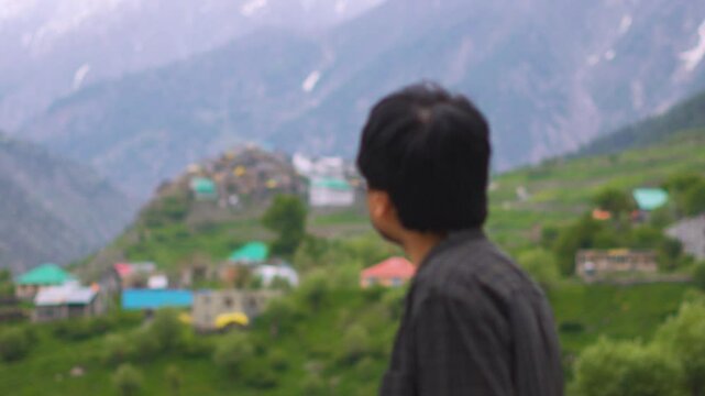 4K Rack focus shot of an Indian man looking at Triloknath village in front of the snowy Himalayan mountain peaks at Pattan valley in Lahaul, Himachal Pradesh, India. Man looking at the village.