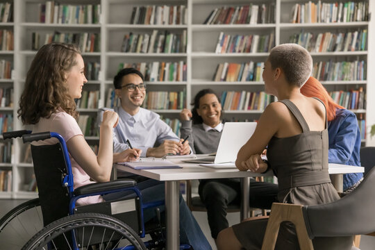 Young female using wheelchair get higher education in friendly inclusive academic group having fun with classmates of diverse ethnicities chat talk discuss funny college university gossips in library