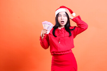 beauty ladies wearing christmas blouse, santa hat, surprised to left, hand to hat and hold cash rupiah money christmas gift, red christmas blouse, isolated on orange background