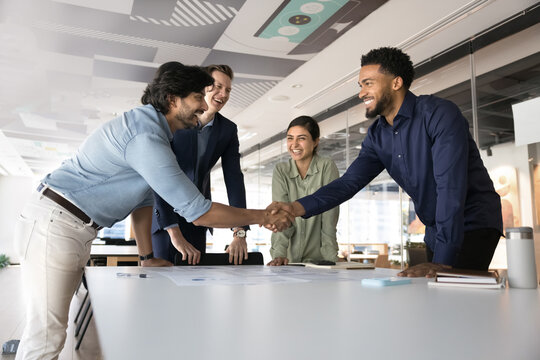 Cheerful positive multiethnic business colleagues celebrating corporate success, shaking hands over meeting table, closing deal, enjoying teamwork, successful startup, partnership, laughing - Powered by Adobe