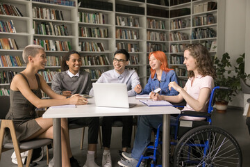 Inclusive education. Happy young female student on wheelchair study at library on training seminar with diverse multiethnic groupmates take notes from video class on notebook work on research in team