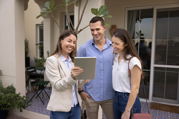 Happy real estate agent woman showing content on tablet to young couple of tenants, renters, house...
