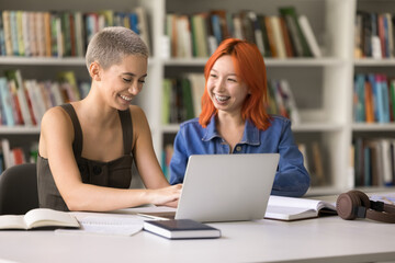 Getting ready for exam. Smiling successful teen girls students classmates of diverse ethnic groups engaged in e-learning use notebook search test answers on scientific website having fun when studying