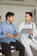 Serious doctor meeting with patient in examination room, making notes, electronic records on...