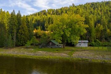 Fototapeta premium Tranquil riverside scene with rustic cabins nestled amongst autumn foliage. Peaceful fall day by the river. Nez Perce-Clearwater National Forest, Syringa, Idaho, USA.