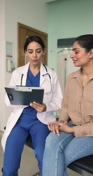 Happy confident female medical specialist talking to Indian woman on appointment in clinic office, asking about healthcare problems, keeping records, smiling, laughing