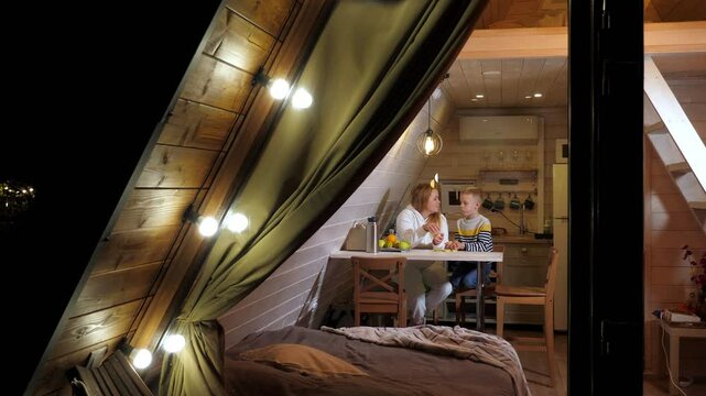 A cheerful mother and son are eating tangerines and having fun in a triangular wooden house, a family vacation in the winter outside the city. A bungalow house in the forest.