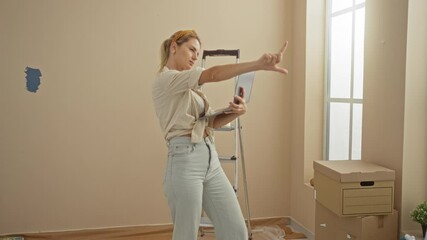 Woman contemplating home renovation using laptop, standing in living room with boxes and ladder, suggesting a move or remodel decision.