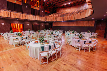 Banquet hall with round tables arranged for an event, decorated with white tablecloths, green napkins, and floral centerpieces on a wooden floor.