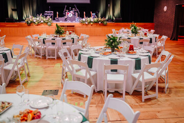 Elegant banquet setup with round tables, white chairs, floral centerpieces, and dishes in a hall with a stage prepared for an event.