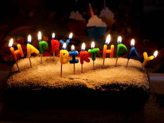 The birthday cake with burning candles in the shape of the lettering “HAPPY BIRTHDAY” is a...