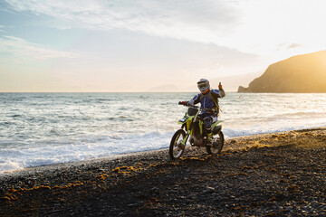 Fototapeta premium Racer shows thumb up at the camera while riding a motorcycle. An enduro rider on an enduro motorcycle on the seashore at the golden sunset. Motocross. Enjoying freedom and an active lifestyle.