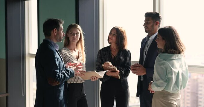 Diverse group of colleagues standing in circle, talking in office hall, discussing work projects, laughing, enjoying professional networking. Positive corporate friends meeting at chatting at window